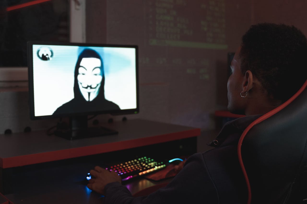 creative-03 Person using computer in dark room viewing hacker wearing a mask on the screen, symbolizing cyber security threats.